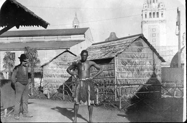 A man stands in traditional African attire, while a man observes.